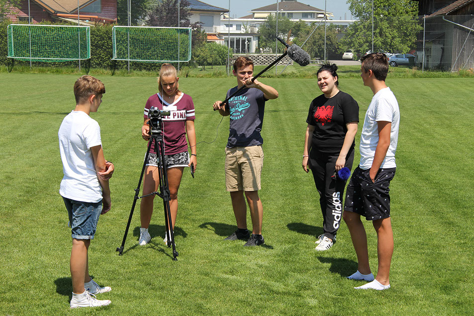 Schülerinnen und Schüler drehen eine Szene auf dem Sportplatz – mit Kamera auf Stativ und Tonangel
