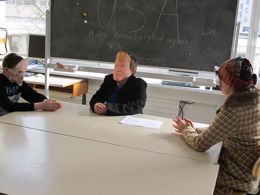 Schüler mit Masken spielen am Tisch eine Szene, an der Wandtafel steht «USA»