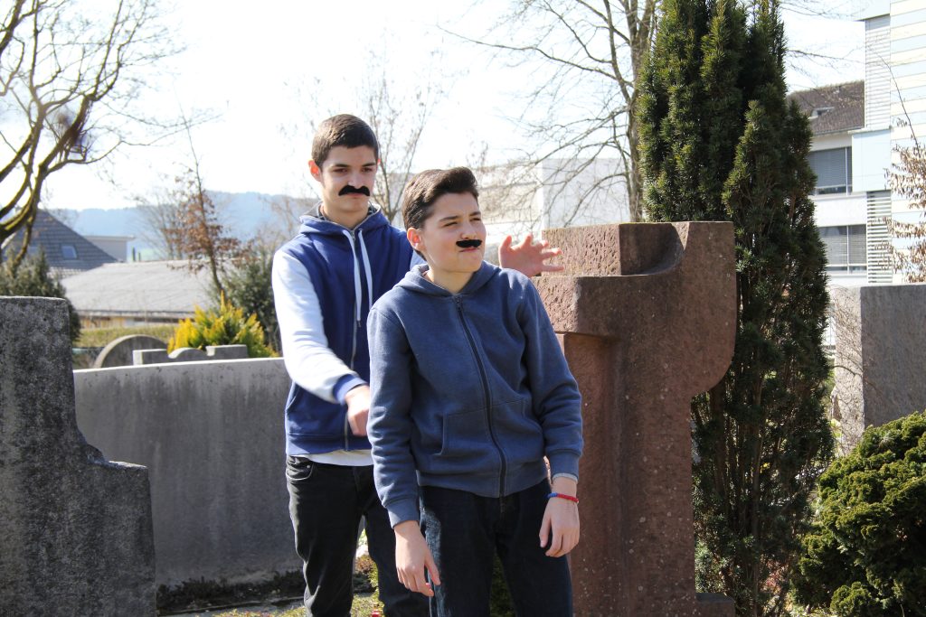 Zwei Schüler mit aufgeklebtem Schnauz spielen eine Szene auf dem Friedhof