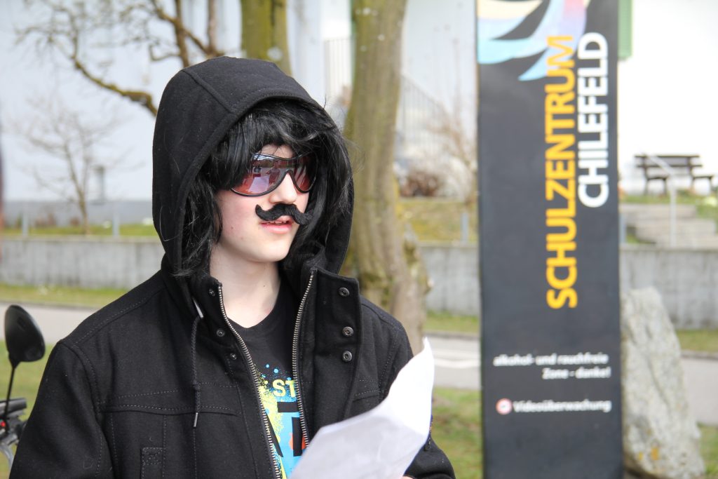 Verkleideter Schüler mit Perücke, Sonnenbrille und Schnauz liest vor dem Schulzentrum Chilefeld aus dem Drehbuch
