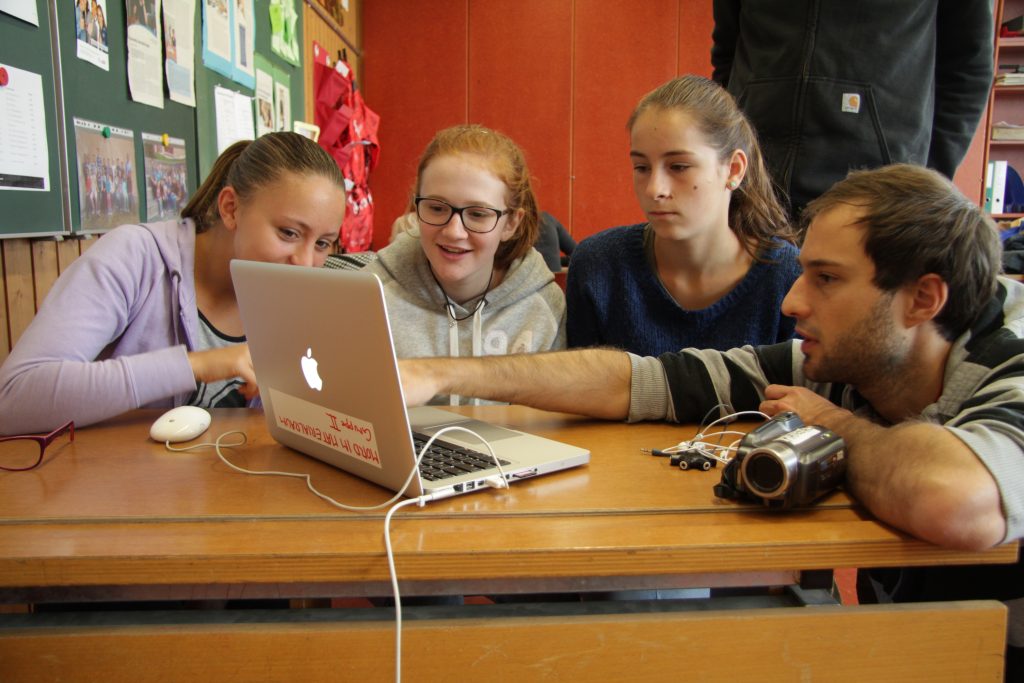 Kursleiter zeigt drei Schülerinnen am Laptop, wie der Filmschnitt funktioniert
